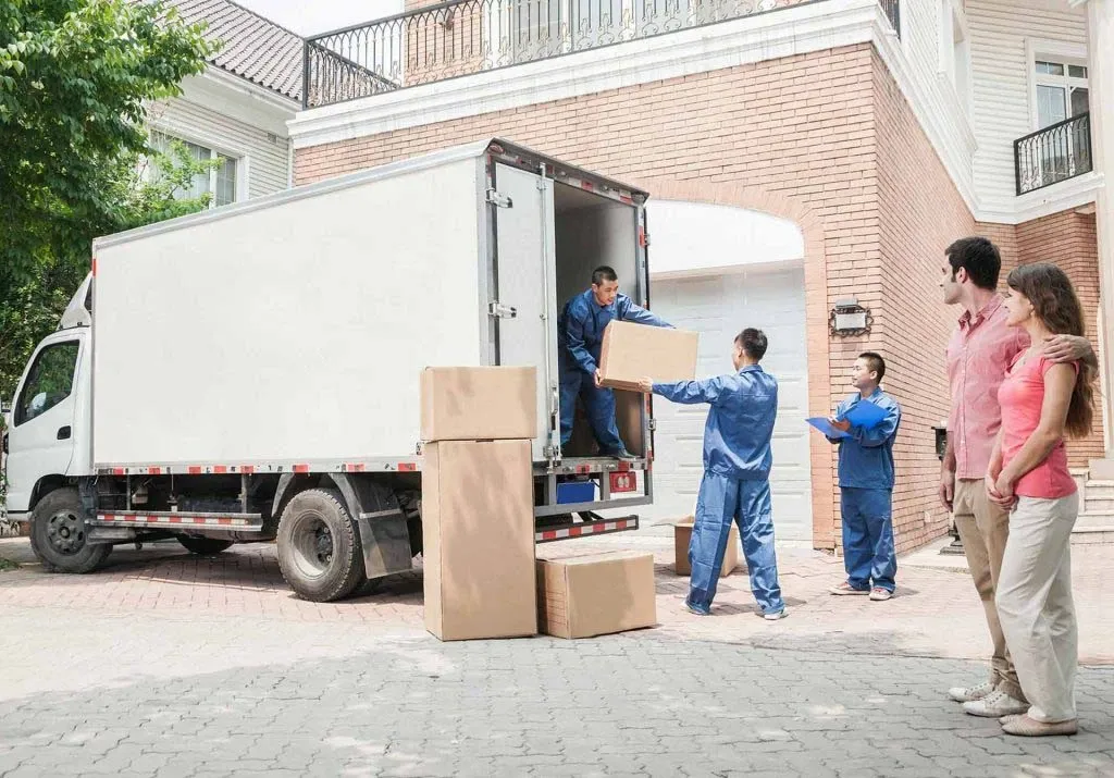 Removalists unloading cartons from a truck while a couple looks on outside their home